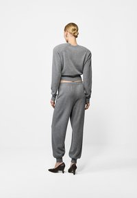 Naine seisab seljaga pöördunu, seljas hall pikaaerderega särk ja sobivad püksid, jalas mustad kontsakingad, valgel taustal.