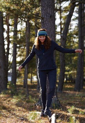 Zwarte fleece jack, marineblauwe shirt, zwarte broek en donkere schoenen. Vrouw in een bos, in een groen-blauwe gebreide hoofdband, in balans op een boomstronk.