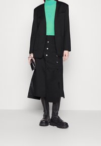Černý blazer přes zelený žebrovaný rolák, doplněný černou sukní na knoflíky a masivními černými botami. Zobrazeny texturované materiály.