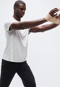 Άνδρας που φοράει μπεζ στηρίγματα καρπού τεντώνει τα χέρια μπροστά, ντυμένος με ανοιχτό γκρι πουκάμισο και μαύρο παντελόνι, μπροστά σε λευκό φόντο.