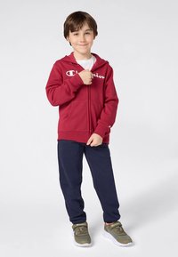 Sweat à capuche rouge zippé avec logo blanc, associé à un pantalon bleu marine et des baskets grises. Tissu lisse, coupe décontractée, poches avant et capuche ajustable.