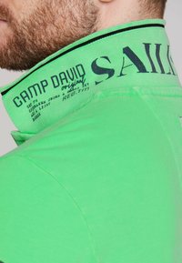 Zielona koszulka polo z wysokim kołnierzem, ozdobiona czarnym tekstem i grafiką. Gładka tekstura z kontrastującymi detalami wzdłuż kołnierza.