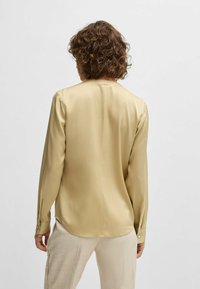 Satin langemouwen overhemd in lichtgeel met een rechte snit, achterplooien en knoopmanchetten. Gladde textuur, subtiele glans en een losse pasvorm.