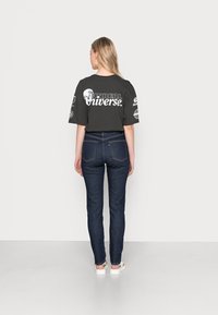 T-shirt gráfica preta com o texto "UNREAL Universe" e várias estampas, combinada com jeans escuros de cintura alta. Sapatos brancos, fundo simples.
