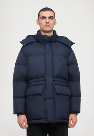 Marineblauwe pufferjack met een hoge kraag, ritssluiting aan de voorkant, twee grote voorkeurs en een verstelbaar trekkoord in de taille. Gladde textuur.