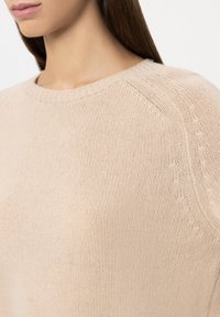 Beige knitted sweater with a crew neckline, smooth texture, and detailed stitching along the shoulder seams. Close-up view.