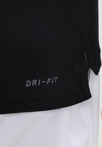 Camiseta negra Dri-Fit, con tejido elegante, presenta un logo gris en el dobladillo, cortada con aberturas laterales, textura suave, adecuada para ropa deportiva.