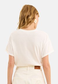 Camiseta de algodón blanca con un corte relajado y mangas enrolladas, con un sutil detalle de logo en la parte posterior, combinada con pantalones de color claro.
