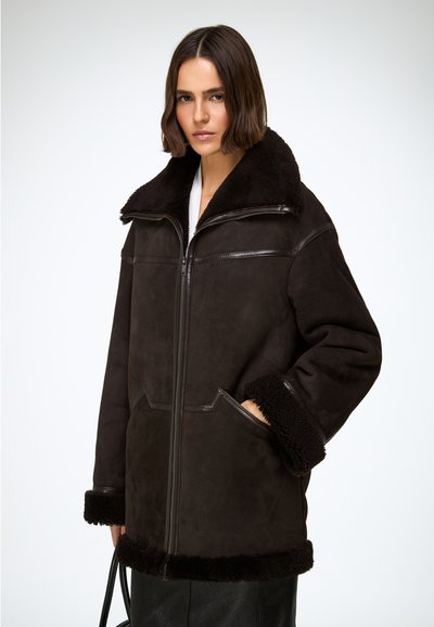 Veste en daim marron avec col et poignets en peau lainée. Elle comprend une fermeture éclair à l'avant, des poches latérales et une texture douce.