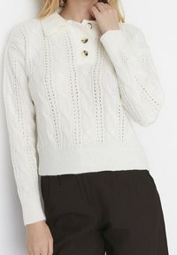 Weißer, kabelgestrickter Pullover mit drei goldenen Knöpfen am Kragen, getragen zu schwarzen Hosen, Hand des Models ruht in Nackennähe.