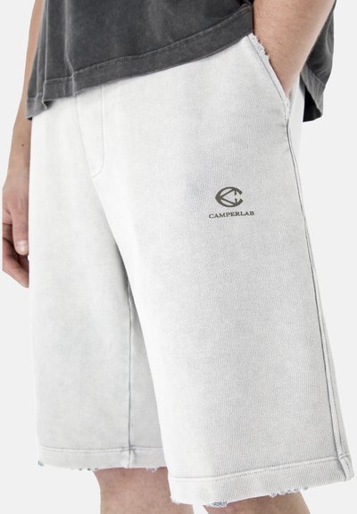 Homme portant un short gris clair texturé avec le logo Camperlab sur la cuisse, main dans la poche, associé à une chemise gris foncé.