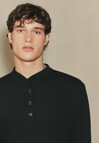 Schwarzes, geripptes Langarmoberteil mit einem geknöpften Henley-Ausschnitt, das eine glatte Textur und vier dunkle Knöpfe auf der Vorderseite hat.