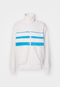 Bílá zipová bunda s texturovaným vzorem, která má tři horizontální modré pruhy přes hruď. Elastické manžety a lem.