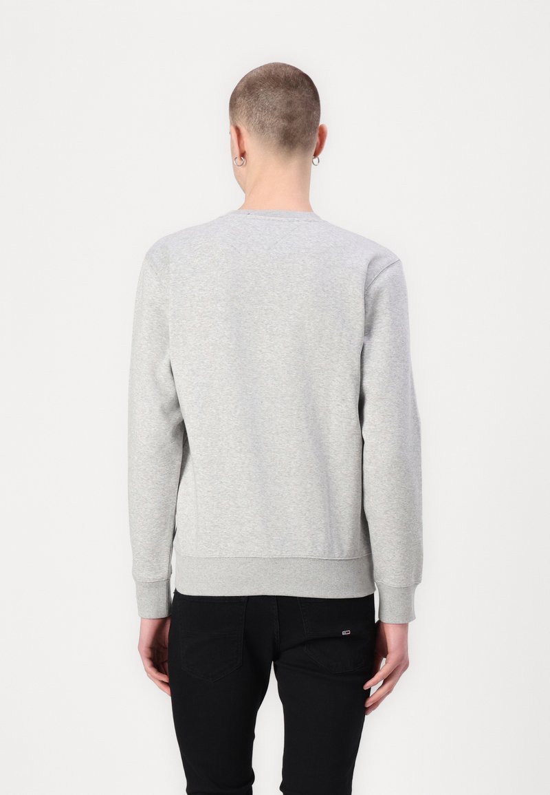 Osoba z krótkimi włosami i kolczykami w kształcie koła, ubrana w jasnoszary długi sweter i czarne dżinsy, zwrócona plecami na białym tle.