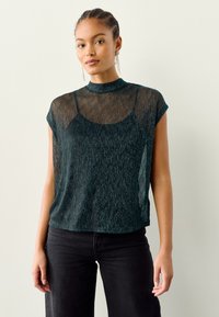 Majica tamno tirkizne boje s kratkim rukavima, visokim izrezom i prozirnom teksturom, slojevito nošena preko jednostavne osnove. Ima suptilan prugasti uzorak.