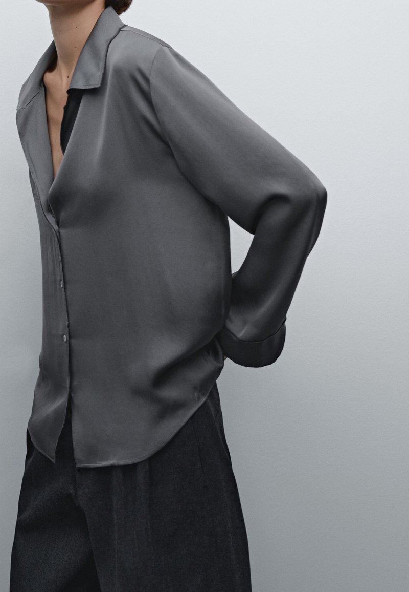 Personne portant une chemise ample à manches longues gris foncé boutonnée et un pantalon foncé, debout avec une main dans le dos sur un fond uni.
