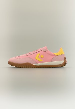 Baskets basses roses avec semelle en caoutchouc, logo étoile jaune sur le côté et languette jaune au talon sur un fond neutre.