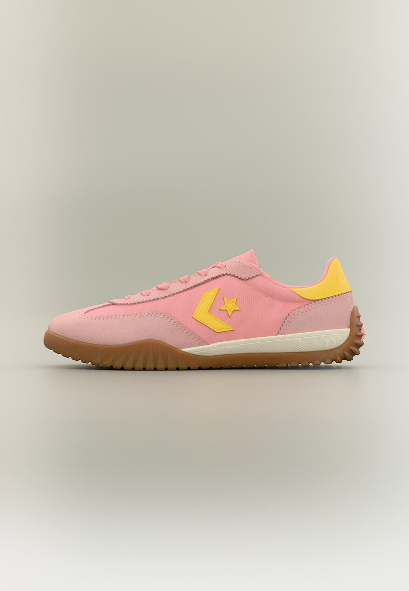 Χαμηλού προφίλ ροζ αθλητικό παπούτσι με σόλα από καουτσούκ, κίτρινο λογότυπο αστεριού στο πλάι και κίτρινη ταμπέλα στη φτέρνα σε ουδέτερο φόντο.
