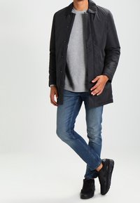 Musta kevyt takki, jossa on terävä kaulus ja napitus, puettu harmaan collegepaidan päälle. Siniset farkut ja mustat lenkkarit täydentävät asun.