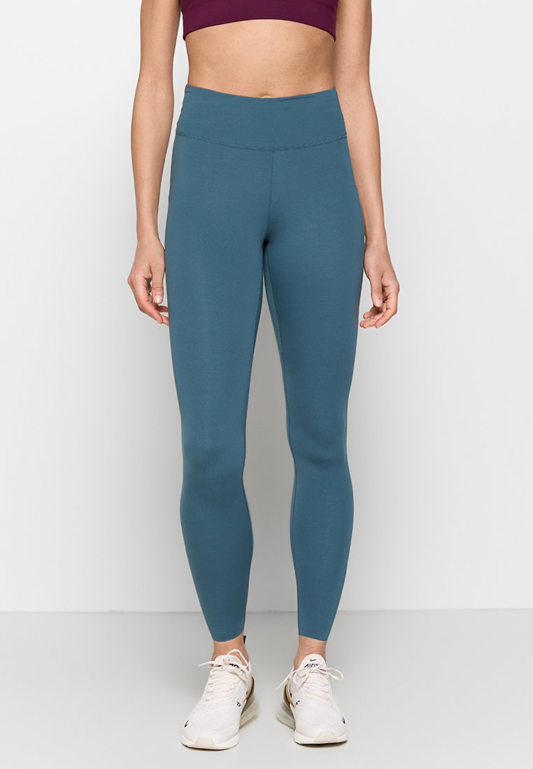 Leggings de color teal con una cintura alta, textura suave y un diseño ajustado, combinados con zapatillas deportivas blancas, de pie sobre un fondo neutro.