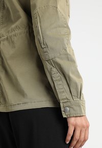 Vaalea khakinvärinen takki, jossa on rento istuvuus, nappikiinnitteiset resorit hihoissa ja pehmustetut kyynärpäiden kohdat, valmistettu sileästä kankaasta.