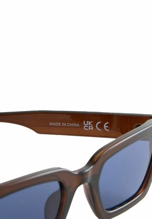 Lunettes de soleil en plastique marron avec une monture angulaire épaisse, dotées de verres teintés foncés et de texte gravé à l'intérieur de la branche.