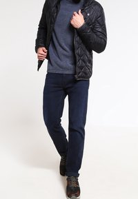 Mann in einer schwarzen Steppjacke, blauer strukturiertem Pullover, dunklen Jeans und sneakers mit Camouflage-Muster, der mit einem Bein nach vorne läuft.