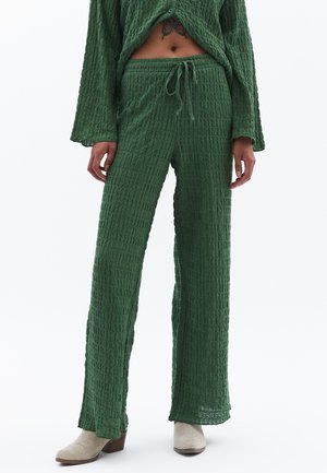 Grünes, strukturiertes Lounge-Set mit einem locker sitzenden Oberteil mit langen Ärmeln und Hose mit Kordelzug, beide aus einem zerknitterten Stoff. Akzentuiert mit beigen Schuhen.