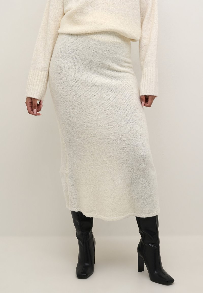 Strikket sæt bestående af sweater og nederdel i off-white, med en blød tekstur og ribbede ærmekanter, parret med sorte højhælede støvler.