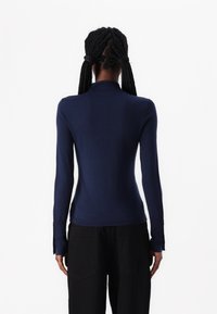 Pull à col roulé à manches longues marine en tissu léger, avec une coupe ajustée et de légentes fentes sur les poignets. Pantalon noir visible.