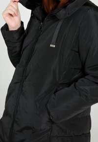 Musta toppatakki, jossa on korkea kaulus, vetoketjukiinnitys ja sivutaskut. Sileä, kiiltävä kangas tikatuilla yksityiskohdilla. Hienovaraiset hopeiset metalliosat.