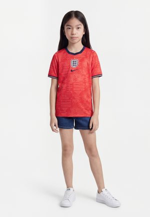 Μικρό κορίτσι που στέκεται φορώντας κόκκινη φανέλα ποδοσφαίρου της Αγγλίας, σκούρο μπλε τζιν σορτς και λευκά αθλητικά παπούτσια σε λευκό φόντο.