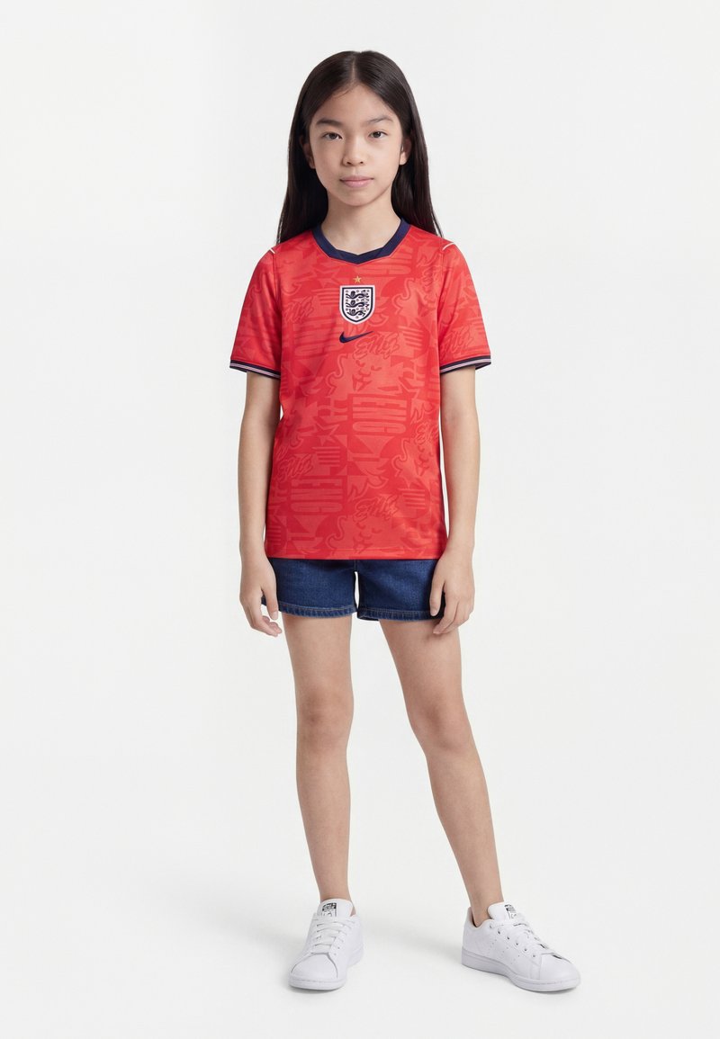 Μικρό κορίτσι που στέκεται φορώντας κόκκινη φανέλα ποδοσφαίρου της Αγγλίας, σκούρο μπλε τζιν σορτς και λευκά αθλητικά παπούτσια σε λευκό φόντο.