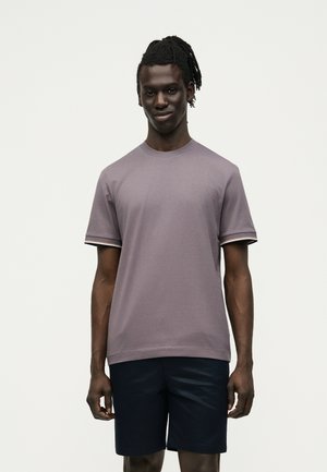 Νεαρός άνδρας με σκούρο δέρμα και πλεξούδες φοράει μια μοβ κοντομάνικη μπλούζα και σκούρα σορτς, στέκεται μπροστά από ένα απλό φωτεινό φόντο.