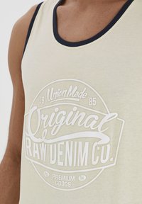 Lys beige tanktop med marineblå trim har et fremtrædende hvidt print, der siger "Original Raw Denim Co.", inklusive detaljer som "Union Made 1985."