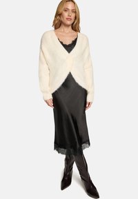 Elfenbeinfarbener, flauschiger Cardigan mit langen Ärmeln, über einem schwarzen Satin-Kleid mit Spitzenbesatz am Saum getragen, kombiniert mit braunen kniehohen Stiefeln.