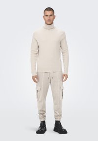Beige gerippter Rollkragenpullover kombiniert mit hellbeigen Cargo-Jogginghosen mit seitlichen Taschen, elastischen Bündchen und schwarzen Ankle Boots.