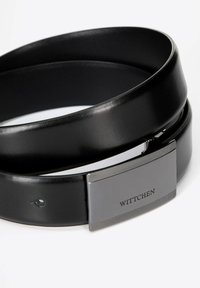 Zwarte leren riem opgerold met een rechthoekige metalen gesp, gegraveerd met "WITTCHEN" op een witte achtergrond.