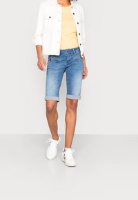 Vaaleansiniset farkkushortsit, joissa on kierretty helma, yhdistettynä keltaiseen puseroon ja valkoiseen takkiin, sekä valkoiset tennarit.