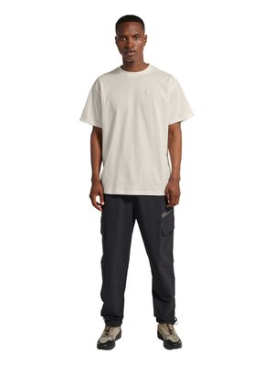 Mand stående med fronten mod kameraet iført en off-white kortærmet skjorte, sorte cargo bukser med tekst og beige chunky sneakers.