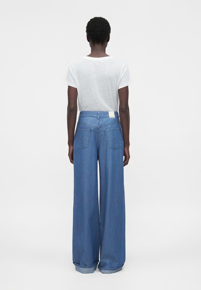 Άτομο με την πλάτη προς την κάμερα, φοράει λευκή μπλούζα και ψηλόμεσα, φαρδιά μπλε τζιν με γυρισμένα ρεβέρ, σε απλό φόντο.