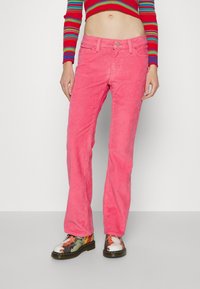 Pantalon en velours côtelé rose à jambes droites avec une finition texturée, design à cinq poches, fermeture à bouton sur le devant, porté avec des bottes à motifs.