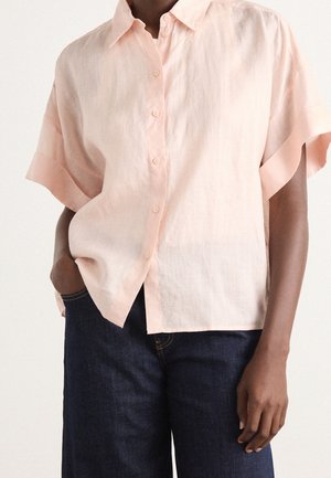 Person trägt ein locker sitzendes, hellrosa kurzärmeliges Hemd mit Knöpfen und hat eine Hand in der Hosentasche einer dunkelblauen Jeans.