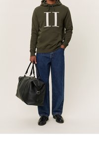 Olivgrön hoodie med vit grafik, blå vida jeans, svart läder duffelväska och svarta skor med texturerade snören.