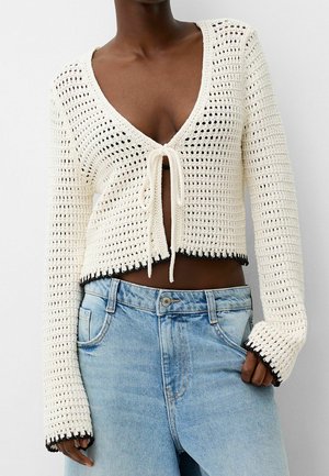 Personne portant un cardigan en crochet blanc à manches longues noué devant avec un bord noir, associé à un jean taille haute bleu clair.
