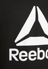 Černé tričko vyrobené z bavlny, které má velké bílé logo s trojúhelníkovým designem a názvem značky "Reebok" v tučných písmech pod ním.