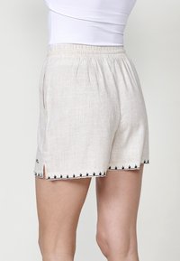 Shorts en lin beige clair avec une ceinture élastique, des poches latérales et des accents brodés noirs le long de l'ourlet et des détails de fente latérale.