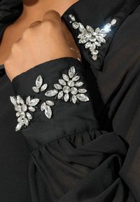 Blusa negra con un cuello estructurado, adornada con strass transparentes en formas de flores y hojas. Presenta una tela suave y texturizada.
