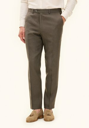 Men’s tailored olive-green dress pants paired with beige tassel loafers and a white buttoned shirt, hands casually in pockets.