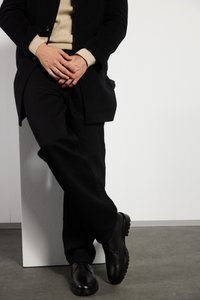 Zapatos de cuero negros con suelas resistentes, combinados con pantalones negros y un suéter beige, apoyados contra un pilar blanco texturizado.